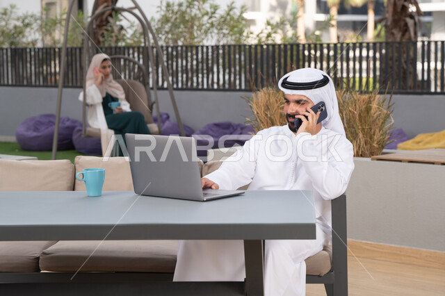 قضاء وقت ممتع فى الخارج ، الاستمتاع بالمشروبات الساخنه ،استخدم جهاز الحاسوب المحمول لانجاز المشاريع والمهام في الكافيه ، مفهوم التكنولوجيا وإستخدام تقنيات حديثة ،شاب عربي خليجي إماراتي يرتدي الكندورة والغترة البيضاء يجلس في المقهى ، تجلس فى الخلف سيدة اماراتيه تجري مكالمه هاتفيه
