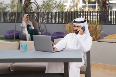 قضاء وقت ممتع فى الخارج ، الاستمتاع بالمشروبات الساخنه ،استخدم جهاز الحاسوب المحمول لانجاز المشاريع والمهام في الكافيه ، مفهوم التكنولوجيا وإستخدام تقنيات حديثة ،شاب عربي خليجي إماراتي يرتدي الكندورة والغترة البيضاء يجلس في المقهى ، تجلس فى الخلف سيدة اماراتيه تجري مكالمه هاتفيه