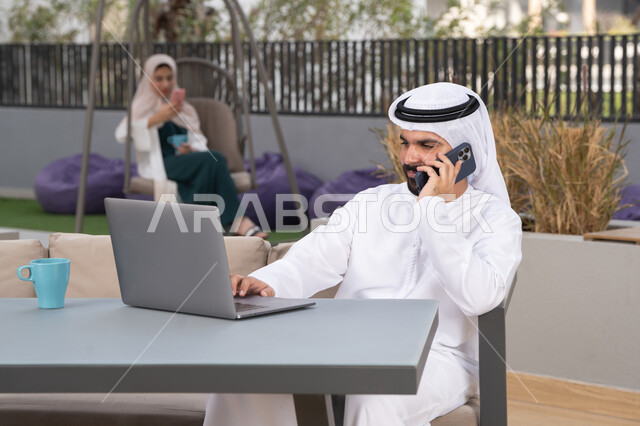 الاستمتاع بالمشروبات الساخنه ،استخدم جهاز الحاسوب المحمول لانجاز المشاريع والمهام في الكافيه ، مفهوم التكنولوجيا وإستخدام تقنيات حديثة ،قضاء وقت ممتع فى الخارج ، شاب عربي خليجي إماراتي يرتدي الكندورة والغترة البيضاء يجلس في المقهى ، تجلس فى الخلف سيدة اماراتيه تجري مكالمه هاتفيه