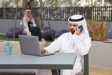 الاستمتاع بالمشروبات الساخنه ،استخدم جهاز الحاسوب المحمول لانجاز المشاريع والمهام في الكافيه ، مفهوم التكنولوجيا وإستخدام تقنيات حديثة ،قضاء وقت ممتع فى الخارج ، شاب عربي خليجي إماراتي يرتدي الكندورة والغترة البيضاء يجلس في المقهى ، تجلس فى الخلف سيدة اماراتيه تجري مكالمه هاتفيه