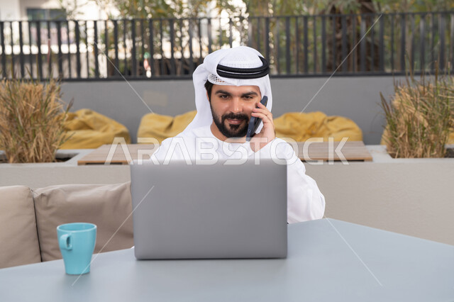 اجراء مكالمه هاتفيه، قضاء وقت ممتع فى الخارج ، الاستمتاع بالمشروبات الساخنه ، شاب عربي خليجي إماراتي يرتدي الكندورة والغترة البيضاء يجلس في المقهى ، استخدم جهاز الحاسوب المحمول لانجاز المشاريع والمهام ، مفهوم التكنولوجيا وإستخدام تقنيات حديثة