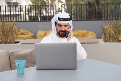 Making a phone call, having a good time outside, enjoying a hot drink, a young Emirati Gulf Arab man wearing a white kandura and ghutra sitting in a cafe, using a laptop to complete projects and tasks, the concept of technology and the use of modern techniques