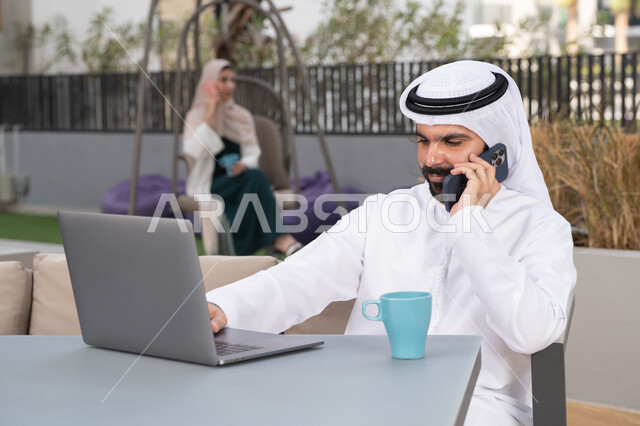 مفهوم التكنولوجيا وإستخدام تقنيات حديثة ، الاستمتاع بالمشروبات الساخنه ،استخدم جهاز الحاسوب المحمول لانجاز المشاريع والمهام في الكافيه ، قضاء وقت ممتع فى الخارج ، شاب عربي خليجي إماراتي يرتدي الكندورة والغترة البيضاء يجلس في المقهى ، تجلس فى الخلف سيدة اماراتيه تجري مكالمه هاتفيه