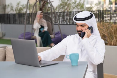 The concept of technology and the use of modern techniques, enjoying hot drinks, using the laptop to complete projects and tasks in the cafe, spending a pleasant time outside, a young Emirati Gulf Arab man wearing a white kandura and ghutra sitting in the cafe, an Emirati woman sitting in the back making a phone call