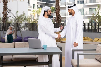 Eid customs in Emirati society, spending enjoyable times with friends, a classy coffee shop, friendships, acquaintances, discussions and dialogues, two Emirati Gulf Arab men wearing kanduras and ghutras shaking hands warmly, happy holidays and occasions