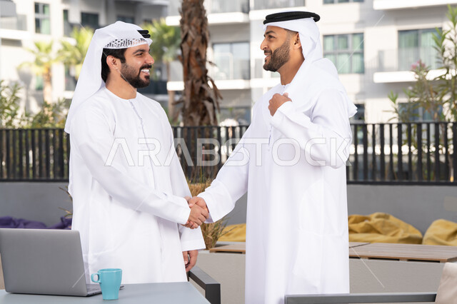 Spending a good time with friends, a classy coffee shop, friendships, acquaintances, discussions and dialogues, Eid customs in the Emirati society, two Emirati Gulf Arab men wearing kanduras and ghutras shaking hands warmly, happy holidays and occasions