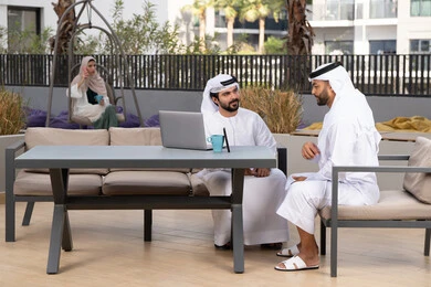 الاستمتاع بالاجواء السعيده ، اجراء مكالمة فيديو ، استخدام الحاسوب المحمول ،دمج التقنيات الحديثه فى الحياه اليومية ، كوفي شوب راقي بالامارات ، صديقان عربيان خليجيان اماراتيان يرتديان الكندورة والغترة البيضاء، يتبادلان الحديث بتعابير السرور ، تجلس فى الخلف سيدة اماراتيه