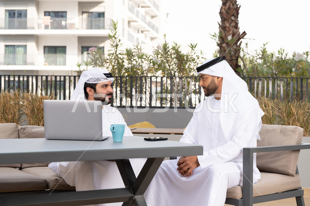 استخدام الحاسوب المحمول ،الاستمتاع بالاجواء السعيده ، اجراء مكالمة فيديو ، دمج التقنيات الحديثه فى الحياه اليومية ، كوفي شوب راقي بالامارات ، صديقان عربيان خليجيان اماراتيان يرتديان الكندورة والغترة البيضاء، يتبادلان الحديث بتعابير السرور