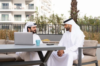 Using a laptop, enjoying the happy atmosphere, making a video call, integrating modern technology into daily life, a sophisticated coffee shop in the Emirates, two Emirati Gulf Arab friends wearing white kandoras and ghutras, exchanging pleasant expressions.