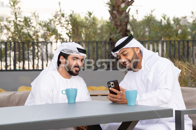 توثيق اللحظات السعيدة، الاستمتاع بالاجواء السعيده ، اجراء مكالمة فيديو ، استخدام الهاتف المحمول ،دمج التقنيات الحديثه فى الحياه اليومية ، كوفي شوب راقي بالامارات ، صديقان عربيان خليجيان اماراتيان يرتديان الكندورة والغترة البيضاء، ينظران بتعابير السرور احدهما يحمل الهاتف المحمول