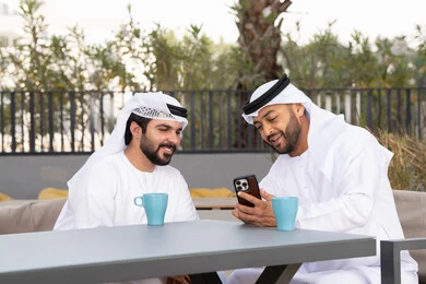 Documenting happy moments, enjoying the happy atmosphere, making a video call, using a mobile phone, integrating modern technologies into daily life, a high-end coffee shop in the Emirates, two Emirati Gulf Arab friends wearing the white kandura and ghutra, looking with happy expressions, one of them holding a mobile phone