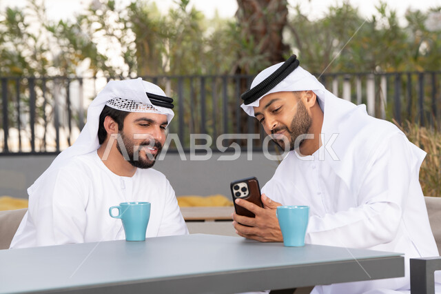 الاستمتاع بالاجواء السعيده ، اجراء مكالمة فيديو ، استخدام الهاتف المحمول ،دمج التقنيات الحديثه فى الحياه اليومية ، كوفي شوب راقي بالامارات ، صديقان عربيان خليجيان اماراتيان يرتديان الكندورة والغترة البيضاء، ينظران بتعابير السرور احدهما يحمل الهاتف المحمول ، توثيق اللحظات السعيدة