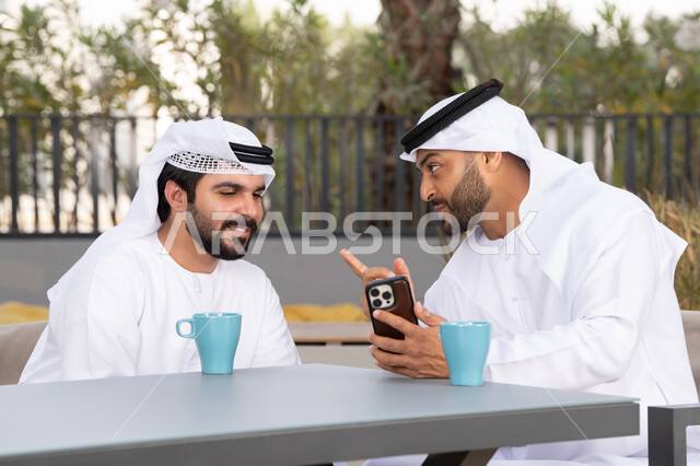 قضاء الاوقات الممتعه ، اجراء مكالمة فيديو ، استخدام الهاتف المحمول ،دمج التقنيات الحديثه فى الحياه اليومية ، كوفي شوب راقي بالامارات ، صديقان عربيان خليجيان اماراتيان يرتديان الكندورة والغترة البيضاء، ينظران بتعابير السرور احدهما يحمل الهاتف المحمول ، توثيق اللحظات السعيدة