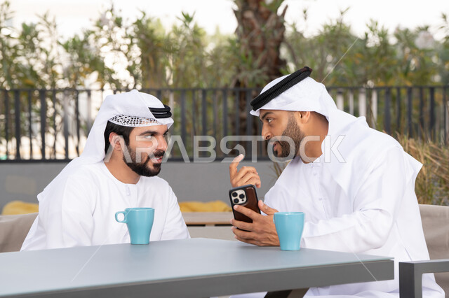 اجراء مكالمة فيديو ، استخدام الهاتف المحمول ،دمج التقنيات الحديثه فى الحياه اليومية ، كوفي شوب راقي بالامارات ، صديقان عربيان خليجيان اماراتيان يرتديان الكندورة والغترة البيضاء، ينظران بتعابير السرور احدهما يحمل الهاتف المحمول ، توثيق اللحظات السعيدة