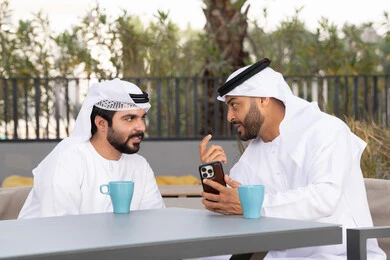 Making a video call, using a mobile phone, integrating modern technologies into daily life, a high-end coffee shop in the Emirates, two Emirati Gulf Arab friends wearing white kanduras and ghutras, looking with happy expressions, one of them holding a mobile phone, documenting happy moments