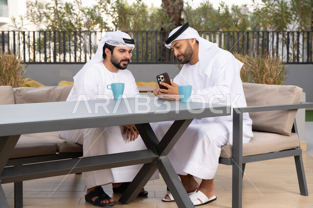 استخدام الهاتف المحمول ،دمج التقنيات الحديثه فى الحياه اليومية ، كوفي شوب راقي بالامارات ، صديقان عربيان خليجيان اماراتيان يرتديان الكندورة والغترة البيضاء، ينظران بتعابير السرور احدهما يحمل الهاتف المحمول ،اجراء مكالمة فيديو ، توثيق اللحظات السعيدة