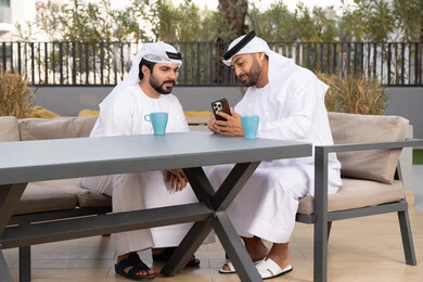 Using a mobile phone, integrating modern technologies into daily life, a high-end coffee shop in the Emirates, two Emirati Gulf Arab friends wearing white kanduras and ghutras, looking with happy expressions, one of them holding a mobile phone, making a video call, documenting happy moments