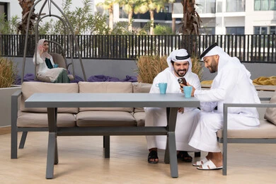 Integrating modern technologies into daily life, a high-end coffee shop in the Emirates, two Emirati Gulf Arab friends wearing white kanduras and ghutras, looking with happy expressions, one of them holding a mobile phone, making a video call, using a mobile phone, documenting happy moments