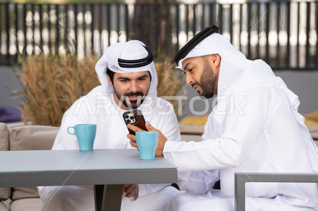 كوفي شوب راقي بالامارات ، صديقان عربيان خليجيان اماراتيان يرتديان الكندورة والغترة البيضاء، ينظران بتعابير السرور احدهما يحمل الهاتف المحمول ،اجراء مكالمة فيديو ، استخدام الهاتف المحمول والتقنيات الحديثه، توثيق اللحظات السعيدة