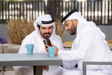 كوفي شوب راقي بالامارات ، صديقان عربيان خليجيان اماراتيان يرتديان الكندورة والغترة البيضاء، ينظران بتعابير السرور احدهما يحمل الهاتف المحمول ،اجراء مكالمة فيديو ، استخدام الهاتف المحمول والتقنيات الحديثه، توثيق اللحظات السعيدة