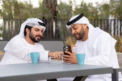 Two Emirati Gulf Arab friends wearing white kandura and ghutra, looking with happy expressions, one of them holding a mobile phone, making a video call, using mobile phones and modern technology, documenting happy moments, upscale coffee shop in the Emirates