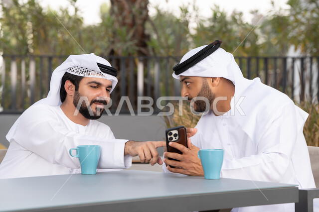اجراء مكالمة فيديو ، استخدام الهاتف المحمول والتقنيات الحديثه، توثيق اللحظات السعيدة،  صديقان عربيان خليجيان اماراتيان يرتديان الكندورة والغترة البيضاء، ينظران بتعابير السرور احدهما يحمل الهاتف المحمول ، كوفي شوب راقي بالامارات