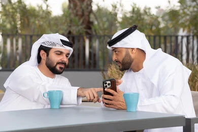 Making a video call, using mobile phones and modern technologies, documenting happy moments, two Emirati Gulf Arab friends wearing white kanduras and ghutras, looking with happy expressions, one of them holding a mobile phone, a high-end coffee shop in the Emirates