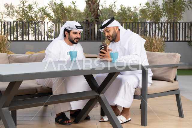 استخدام الهاتف المحمول والتقنيات الحديثه، توثيق اللحظات السعيدة، اجراء مكالمة فيديو ، صديقان عربيان خليجيان اماراتيان يرتديان الكندورة والغترة البيضاء، ينظران بتعابير السرور احدهما يحمل الهاتف المحمول ، كوفي شوب راقي بالامارات