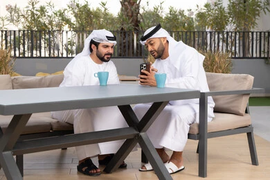استخدام الهاتف المحمول والتقنيات الحديثه، توثيق اللحظات السعيدة، اجراء مكالمة فيديو ، صديقان عربيان خليجيان اماراتيان يرتديان الكندورة والغترة البيضاء، ينظران بتعابير السرور احدهما يحمل الهاتف المحمول ، كوفي شوب راقي بالامارات