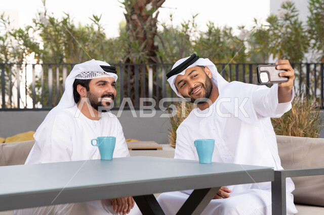 Documenting happy moments, using mobile phones and modern technologies, making a video call, two Emirati Gulf Arab friends wearing white kanduras and ghutras, looking with happy expressions, one of them holding a mobile phone, a high-end coffee shop in the Emirates