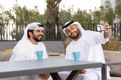 Documenting happy moments, using mobile phones and modern technologies, making a video call, two Emirati Gulf Arab friends wearing white kanduras and ghutras, looking with happy expressions, one of them holding a mobile phone, a high-end coffee shop in the Emirates
