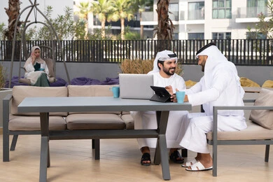 Dialogue and discussion between colleagues, gestures of integration and concentration, a young Emirati Arab man wearing a white kandura and ghutra sitting with his friend in a cafe, enjoying hot drinks, using a tablet and laptop to complete projects and tasks in the cafe, spending quality time with friends