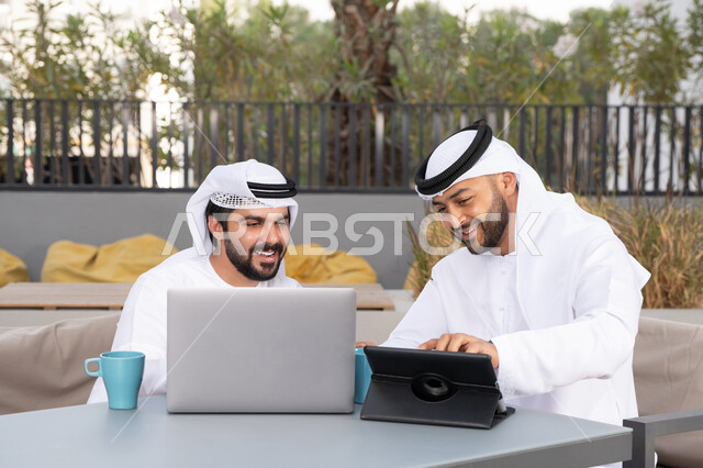ايماءات الاندماج والتركيز ، الحوار والنقاش بين زملاء العمل، شاب عربي خليجي إماراتي يرتدي الكندورة والغترة البيضاء يجلس مع صديقه في المقهى ، الاستمتاع بالمشروبات الساخنه، استخدم جهاز التابلت والحاسوب المحمول لانجاز المشاريع والمهام في الكافيه، قضاء وقت ممتع مع الأصدقاء