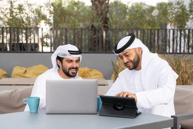 ايماءات الاندماج والتركيز ، الحوار والنقاش بين زملاء العمل، شاب عربي خليجي إماراتي يرتدي الكندورة والغترة البيضاء يجلس مع صديقه في المقهى ، الاستمتاع بالمشروبات الساخنه، استخدم جهاز التابلت والحاسوب المحمول لانجاز المشاريع والمهام في الكافيه، قضاء وقت ممتع مع الأصدقاء