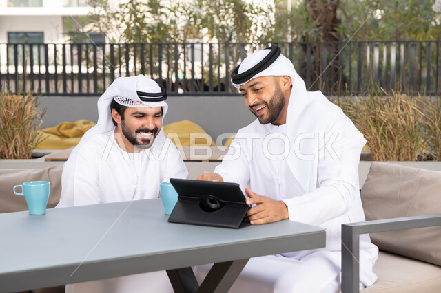 ايماءات الاندماج والتركيز ، الحوار والنقاش بين زملاء العمل، شاب عربي خليجي إماراتي يرتدي الكندورة والغترة البيضاء يجلس مع صديقه في المقهى ، الاستمتاع بالمشروبات الساخنه، استخدم جهاز التابلت لانجاز المشاريع والمهام في الكافيه، قضاء وقت ممتع مع الأصدقاء