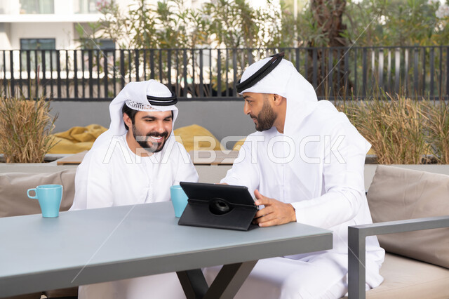 الحوار والنقاش بين زملاء العمل، شاب عربي خليجي إماراتي يرتدي الكندورة والغترة البيضاء يجلس مع صديقه في المقهى ، الاستمتاع بالمشروبات الساخنه، ايماءات الاندماج والتركيز ، استخدم جهاز التابلت لانجاز المشاريع والمهام في الكافيه، قضاء وقت ممتع مع الأصدقاء