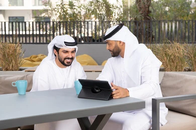 Dialogue and discussion between colleagues, a young Emirati Gulf Arab man wearing a white kandura and ghutra sitting with his friend in a cafe, enjoying hot drinks, gestures of integration and concentration, using the tablet to complete projects and tasks in the cafe, spending quality time with friends