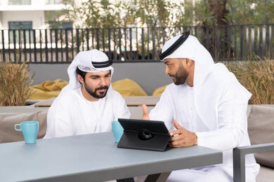 A young Emirati Gulf Arab man wearing a white kandura and ghutra sitting with his friend in a cafe, enjoying hot drinks, gestures of integration and concentration, dialogue and discussion between colleagues, using the tablet to complete projects and tasks in the cafe, spending a fun time with friends