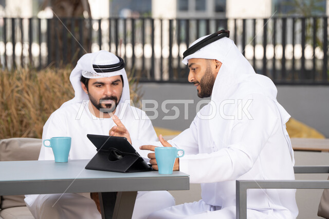 الاستمتاع بالمشروبات الساخنه، ايماءات الاندماج والتركيز ، الحوار والنقاش بين زملاء العمل، استخدم جهاز التابلت لانجاز المشاريع والمهام في الكافيه، قضاء وقت ممتع مع الأصدقاء, شاب عربي خليجي إماراتي يرتدي الكندورة والغترة البيضاء يجلس مع صديقه في المقهى
