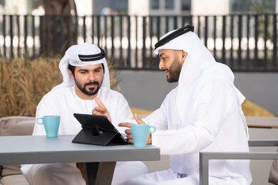 Enjoying hot drinks, gestures of integration and concentration, dialogue and discussion between colleagues, using the tablet to complete projects and tasks in the café, spending quality time with friends, a young Emirati Gulf Arab man wearing a white kandura and ghutra sitting with his friend in the café