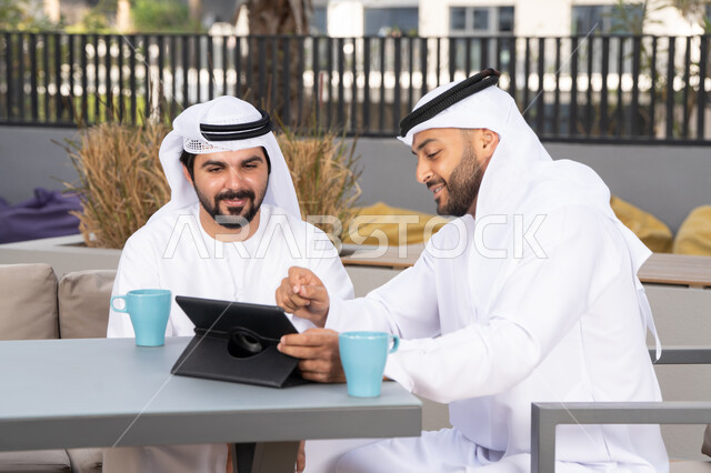 Dialogue and discussion between colleagues, using the tablet to complete projects and tasks in the café, spending quality time with friends, a young Emirati Gulf Arab man wearing a white kandura and ghutra sitting with his friend in the café, gestures of integration and concentration, enjoying hot drinks
