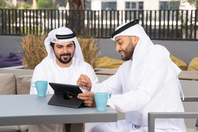 Dialogue and discussion between colleagues, using the tablet to complete projects and tasks in the café, spending quality time with friends, a young Emirati Gulf Arab man wearing a white kandura and ghutra sitting with his friend in the café, gestures of integration and concentration, enjoying hot drinks