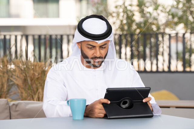 Working remotely, enjoying hot drinks, using modern technology and techniques, a smiling Emirati Gulf Arab man wearing a white kandura and ghutra sitting in an upscale coffee shop in the Emirates, using a tablet to complete projects in the cafe