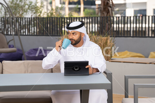 Enjoying hot drinks, using modern technology and techniques, a smiling Emirati Gulf Arab man wearing a white kandura and ghutra sitting in an upscale coffee shop in the Emirates, working remotely, using a tablet to complete projects in the cafe.