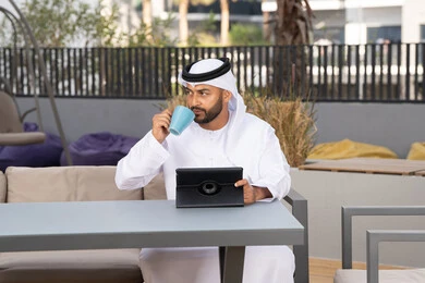 الاستمتاع بالمشروبات الساخنه، استخدام التكنولوجيا والتقنيات الحديثه، رجل عربي خليجي اماراتي مبتسم يرتدي الكندورة والغترة البيضاء يجلس في كوفي شوب راقي بالامارات ، العمل عن بعد، استخدام جهاز التابلت لانجاز المشاريع في المقهي