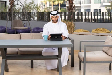 Using modern technology and techniques, a smiling Emirati Gulf Arab man wearing a white kandura and ghutra sitting in an upscale coffee shop in the Emirates, working remotely, enjoying hot drinks, using a tablet to complete projects in the cafe.