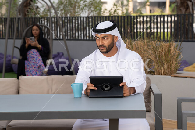 العمل عن بعد، الاستمتاع بالمشروبات الساخنه، اجراء مكالمة هاتفية ،استخدام جهاز التابلت لانجاز المشاريع في المقهى، اجراء مكالمة فيديو عبر الاجهزة التقنية الحديثة، رجل عربي خليجي اماراتي يرتدي الكندورة والغترة البيضاء يجلس في كوفي شوب راقي بالامارات ، تجلس فى الخلف سيدة اماراتية تستخدم الجوال
