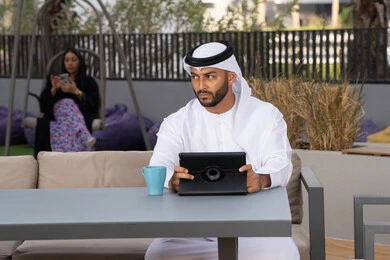 العمل عن بعد، الاستمتاع بالمشروبات الساخنه، اجراء مكالمة هاتفية ،استخدام جهاز التابلت لانجاز المشاريع في المقهى، اجراء مكالمة فيديو عبر الاجهزة التقنية الحديثة، رجل عربي خليجي اماراتي يرتدي الكندورة والغترة البيضاء يجلس في كوفي شوب راقي بالامارات ، تجلس فى الخلف سيدة اماراتية تستخدم الجوال