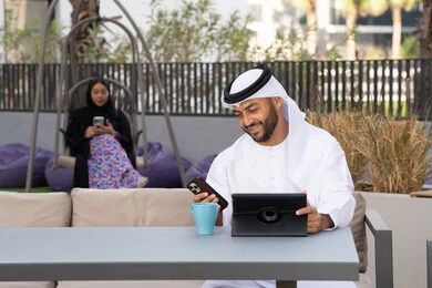 رجل عربي خليجي اماراتي مبتسم يرتدي الكندورة والغترة البيضاء يجلس في كوفي شوب راقي بالامارات ، تجلس فى الخلف سيدة اماراتية تستخدم الجوال، العمل عن بعد، الاستمتاع بالمشروبات الساخنه، اجراء مكالمة هاتفية ،استخدام جهاز التابلت لانجاز المشاريع في المقهى، اجراء مكالمة فيديو عبر الاجهزة التقنية الحديثة