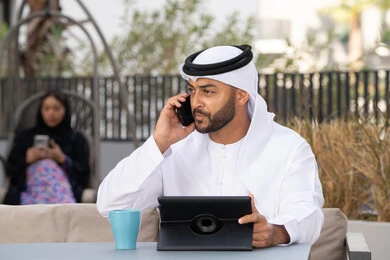 الاستمتاع بالمشروبات الساخنه، اجراء مكالمة هاتفية ،استخدام جهاز التابلت لانجاز المشاريع في المقهى، اجراء مكالمة فيديو عبر الاجهزة التقنية الحديثة، العمل عن بعد، رجل عربي خليجي اماراتي يرتدي الكندورة والغترة البيضاء يجلس في كوفي شوب راقي بالامارات ، تجلس فى الخلف سيدة اماراتية تستخدم الجوال